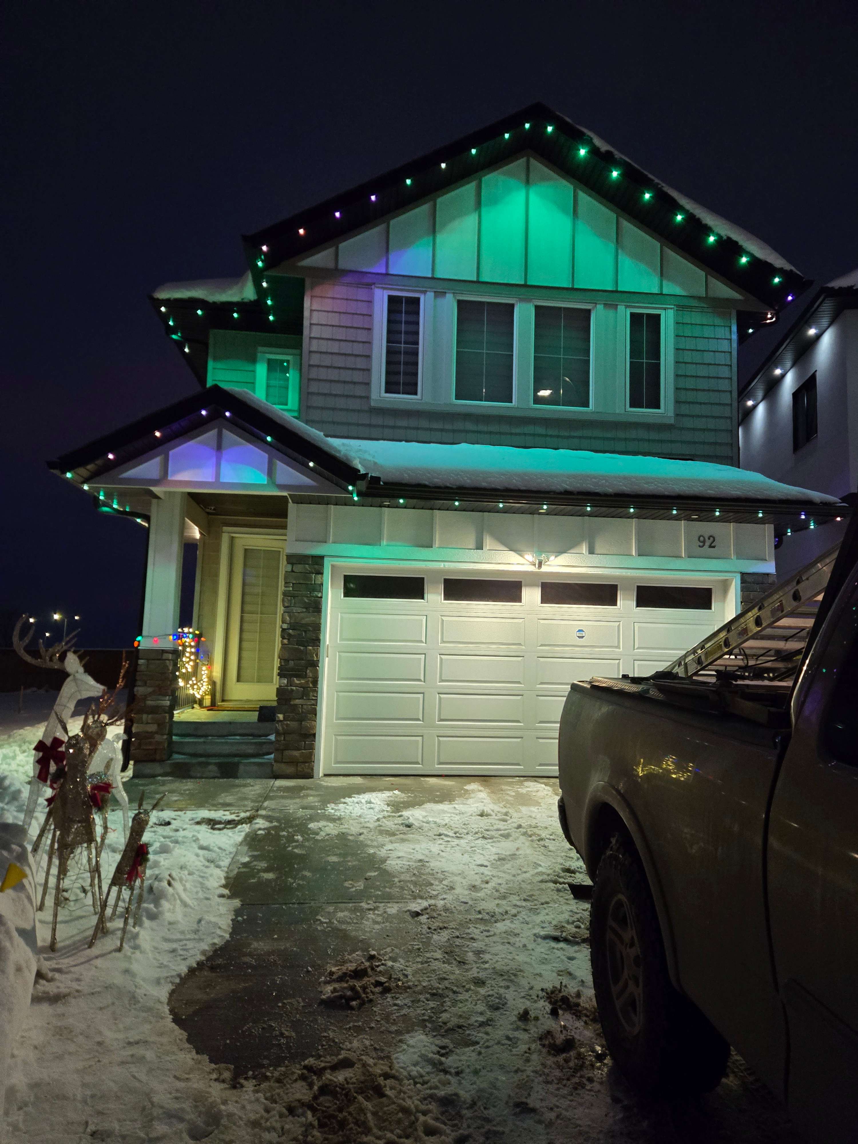 Two-storey home lit green and teal with lit reindeer decorations in the front yard
